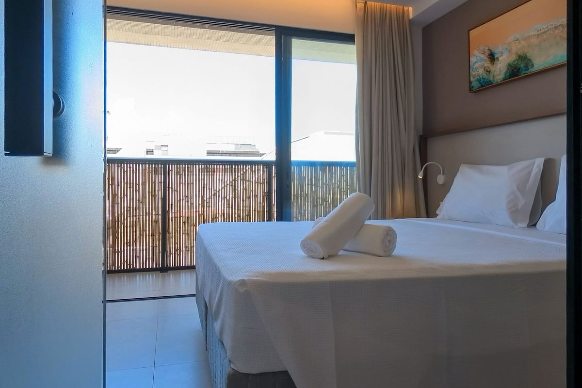 Une chambre d’hôtel avec un lit, une fenêtre avec des rideaux, un tableau au mur et une vue sur les bâtiments environnants.