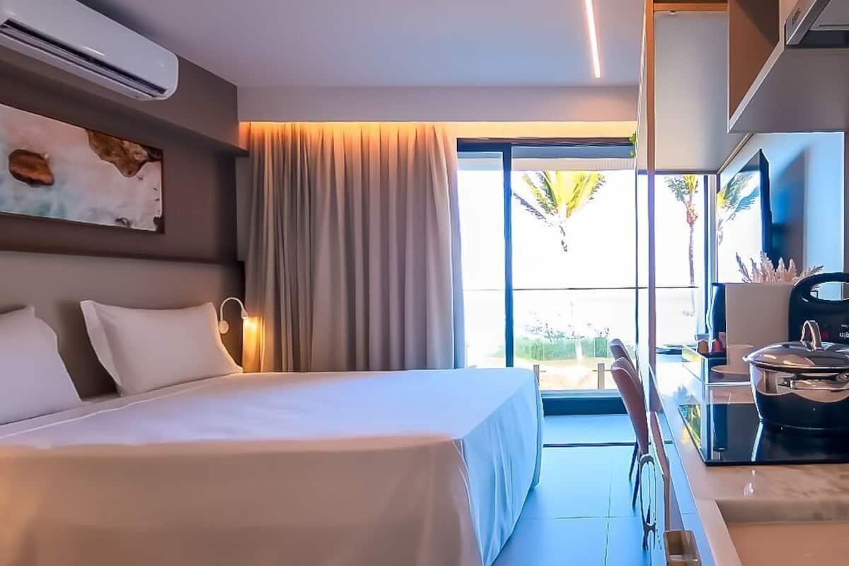 Une chambre d’hôtel moderne dotée d’un grand lit, d’un bureau avec un ordinateur, d’une fenêtre avec des rideaux et offrant une vue sur des palmiers.