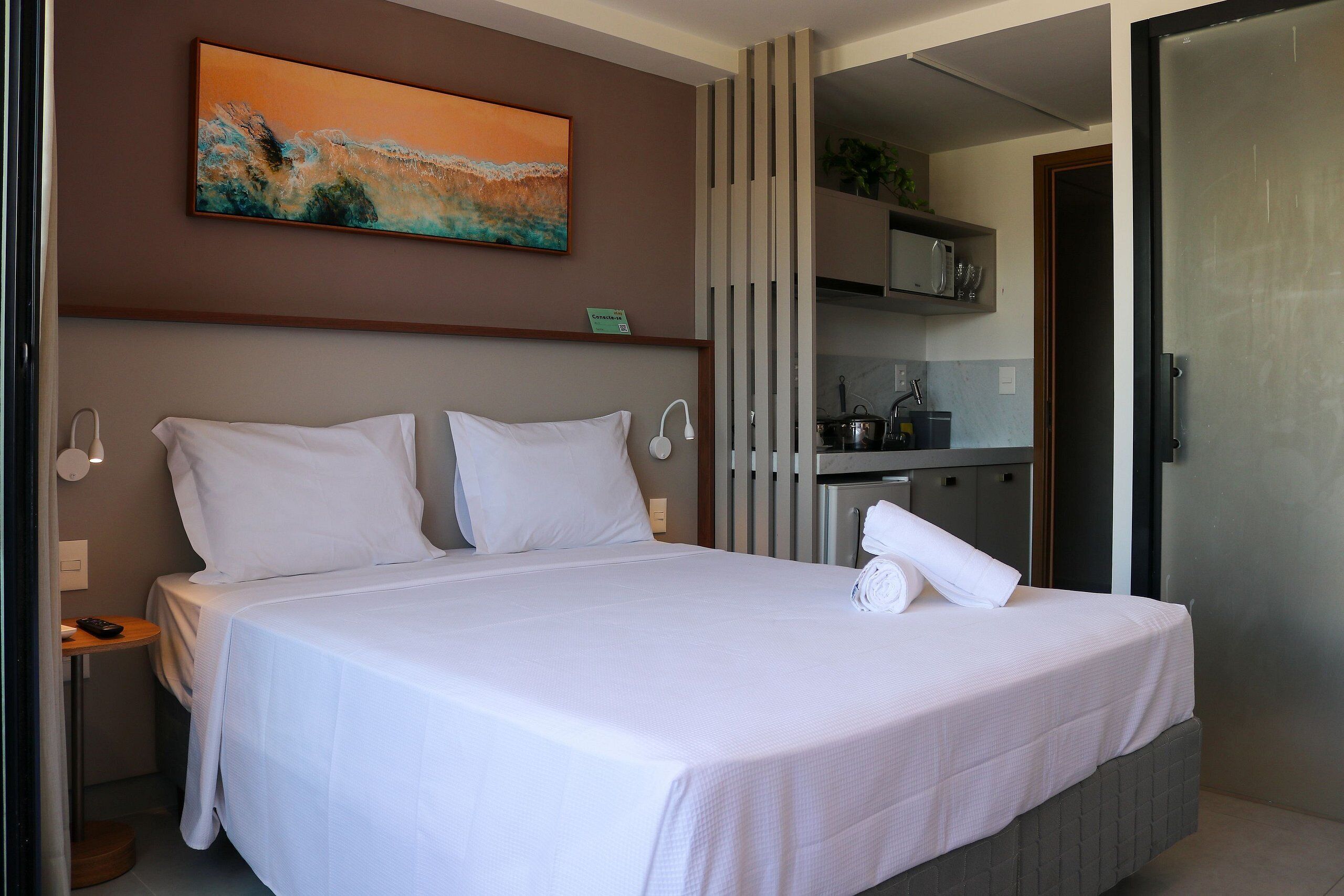 Una cama bien hecha con ropa blanca, una mesita de noche con control remoto y un cuadro sobre la cama.
