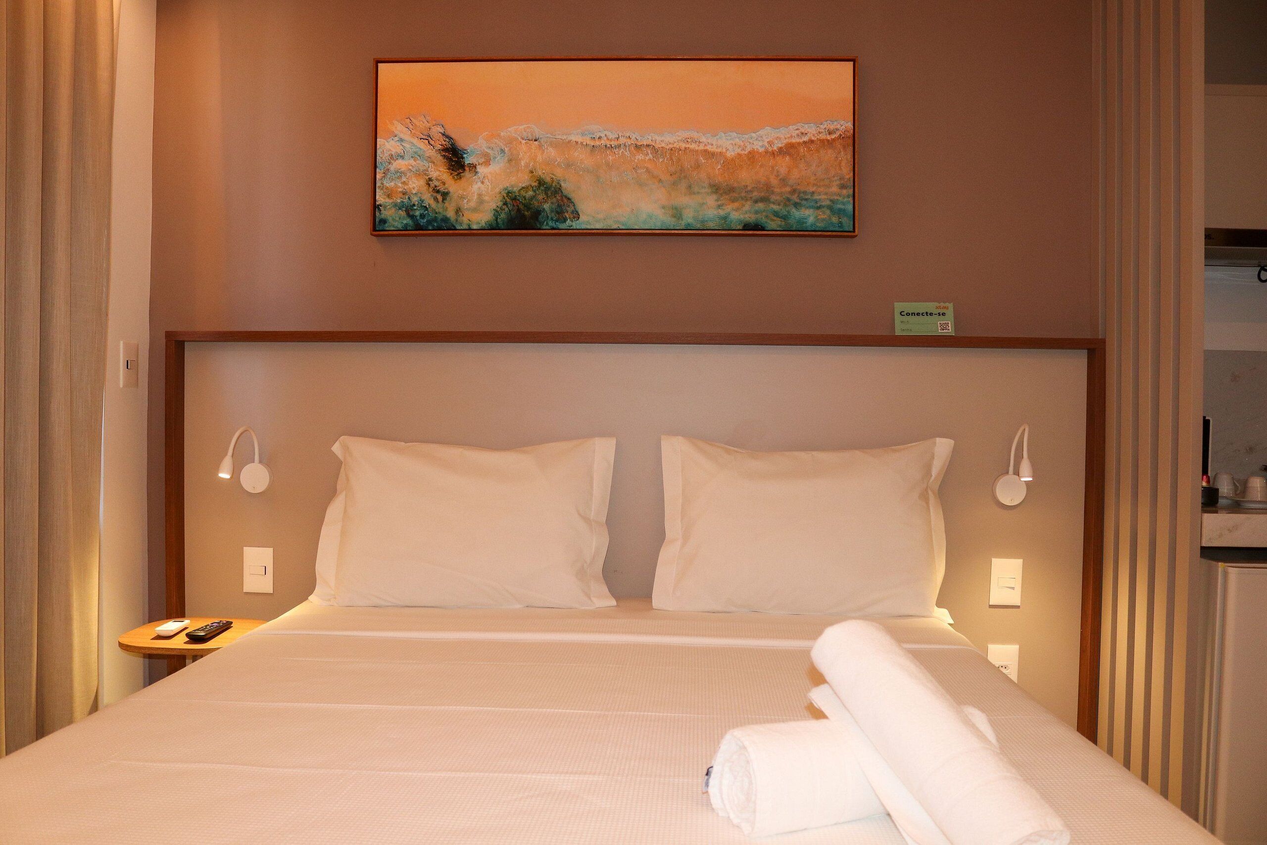 Una cama bien tendida con sábanas blancas, dos almohadas y una toalla enrollada a un lado. Hay un cuadro sobre la cama y una mesita con un control remoto al lado.
