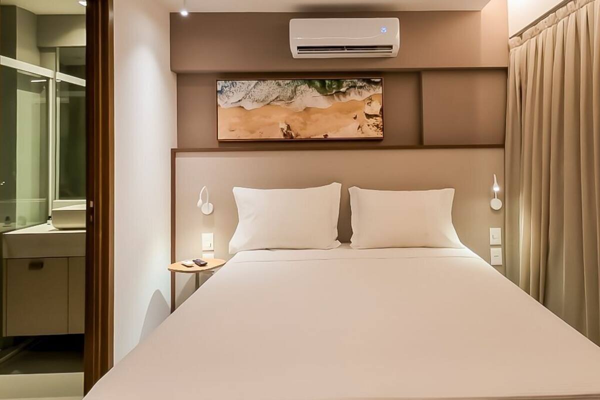 Une chambre d’hôtel moderne avec un grand lit, un appareil de climatisation, un tableau au-dessus du lit et une vue sur la salle de bain à travers une porte en verre.