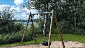 A swing set in a park with a sandy area and a lake in the background. - Gård 2 hus, bastu, internet med fiber. Lugnt (Dannäs)