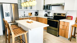Una cocina moderna con armarios blancos, una isla central, taburetes de madera, electrodomésticos de acero inoxidable y un tostador rojo.