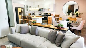 Una sala de estar moderna con un sofá, una mesa de comedor y sillas. Se ve una cocina en el fondo con armarios blancos y electrodomésticos de acero inoxidable.