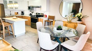 Una cocina moderna con una isla central, armarios blancos, electrodomésticos de acero inoxidable y una mesa de vidrio redonda con cuatro sillas.