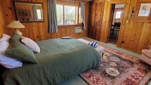 A cozy wooden cabin bedroom with a bed, a small table, a rug, and a window with curtains.