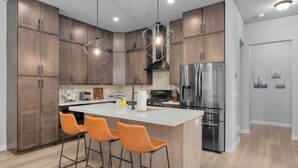 Cozinha moderna com armários de madeira, geladeira de aço inoxidável, ilha central com banquetas e iluminação pendente.