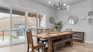 Uma sala de jantar com uma mesa de madeira grande, seis cadeiras, um vaso com plantas, um relógio e quadros nas paredes.
