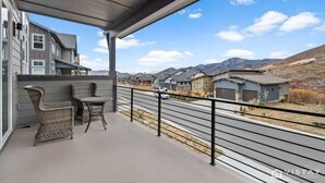 Uma varanda com móveis de vime, grade de metal e vista para uma rua residencial e montanhas.