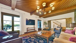 A spacious living room with a wooden ceiling, large windows, a flat-screen TV, and a staircase leading to an upper level. - Villa Encanto- Luxe 6BRs • Courtyard • Game Room (Long Beach)