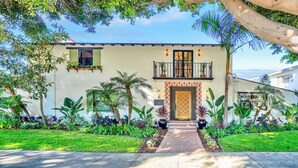 A two-story house with a balcony, a front door with a decorative panel, and a garden with various plants. - Villa Encanto- Luxe 6BRs • Courtyard • Game Room (Long Beach)