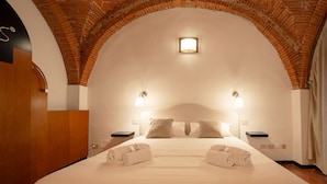 Une chambre d’hôtel avec un grand lit, deux tables de chevet, une petite table d’appoint, un plafond en forme d’arc en briques et une petite fenêtre.