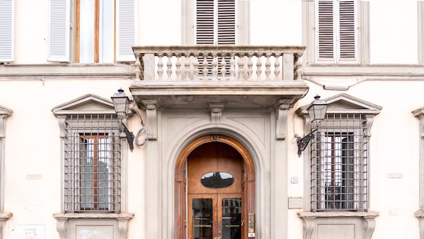 Une façade d’immeuble avec une entrée centrale en arc de cercle, flanquée de deux fenêtres dotées de grilles en métal, et un balcon au-dessus.