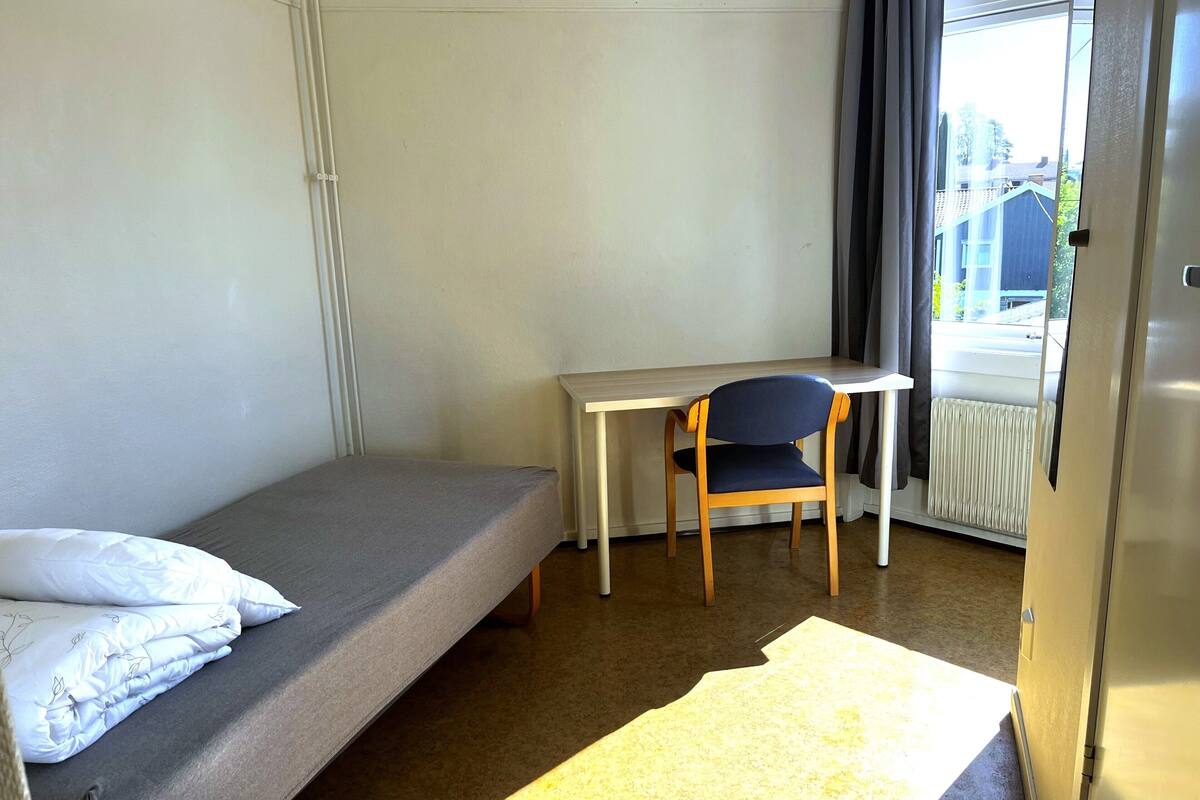 Une petite chambre avec un lit simple, un bureau et une chaise, un radiateur et une fenêtre avec des rideaux.
