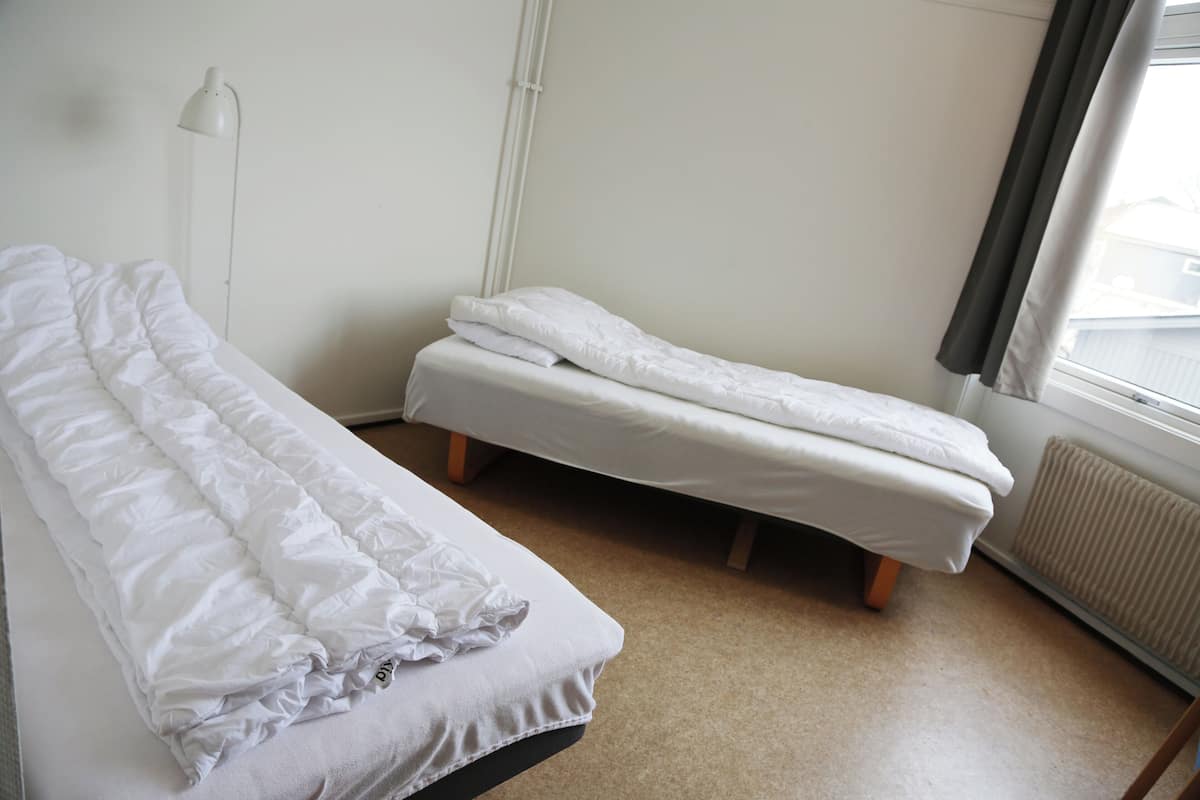 Une petite chambre propre, avec deux lits simples, une table de chevet, une lampe et une fenêtre avec des rideaux.