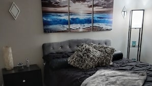 A bedroom with a large bed, a nightstand, a small table, and a wall-mounted shelf.