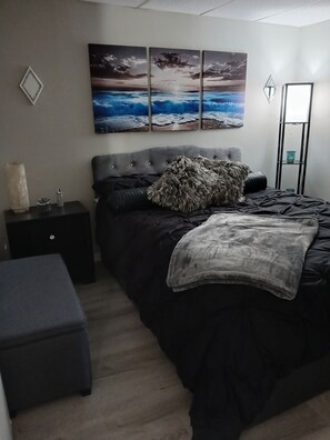 A bedroom with a large bed, a nightstand, a small table, and a wall-mounted shelf.