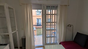 Une pièce comprenant un lit, une table, une armoire et un radiateur. On peut voir, par la fenêtre, un balcon avec une balustrade et un immeuble en arrière-plan.