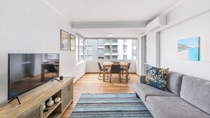 A modern living room with a flat-screen TV, wooden dining table, grey sofa, and large windows offering a view of the city.