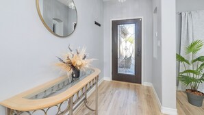 A hallway with a wooden floor, a glass console table with a vase of dried flowers, a round mirror, and a door with a decorative glass panel. - Spacious 4-bedroom house with Pool, Fun BackYard & Amazing Game Room (Bakersfield)