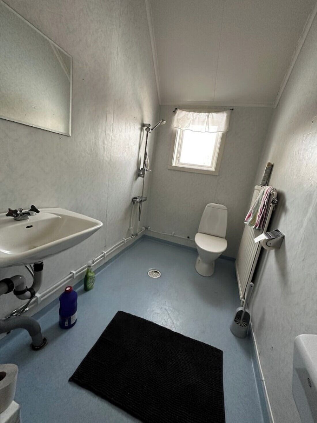 A small, compact bathroom with a white sink, toilet, and shower. A window provides natural light, and a radiator is mounted on the wall.