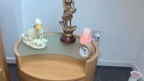 Une table de chevet en bois avec un plateau en verre, une petite statue, une figurine de canard et une lampe de sel.