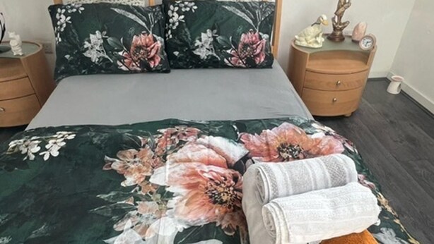 Un lit bien fait, avec une literie à motifs floraux, deux oreillers et une table de chevet sur laquelle se trouvent une lampe et une petite figurine.