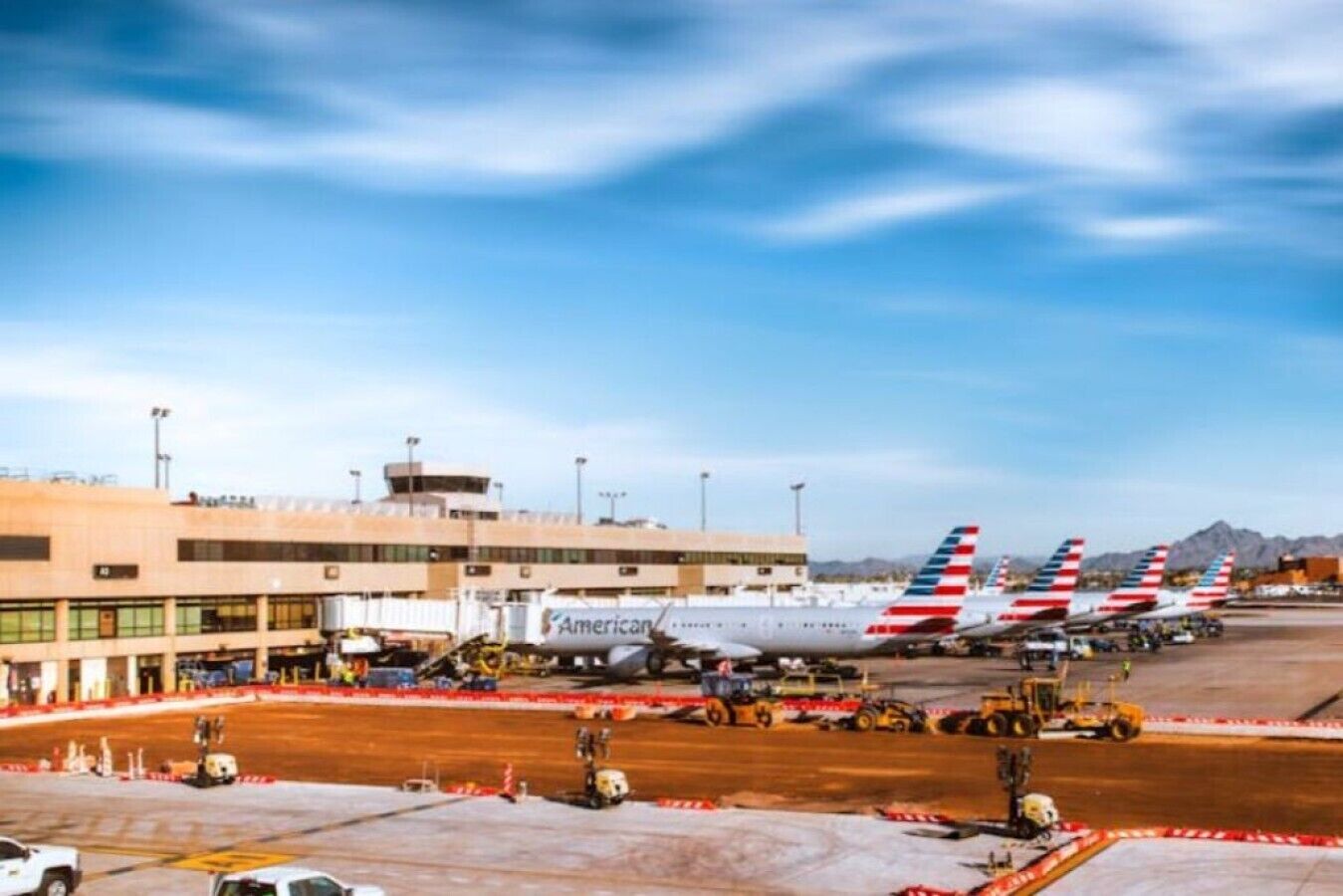 An airport tarmac with multiple American Airlines planes parked, airport buildings, and ground service vehicles.