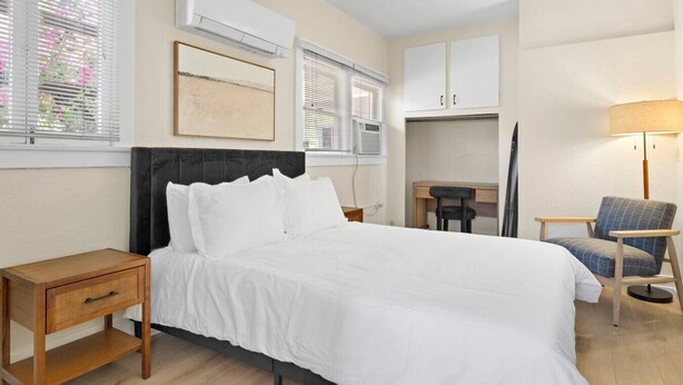 Quarto de hotel com cama, mesas de cabeceira, escrivaninha, cadeira e ar condicionado.