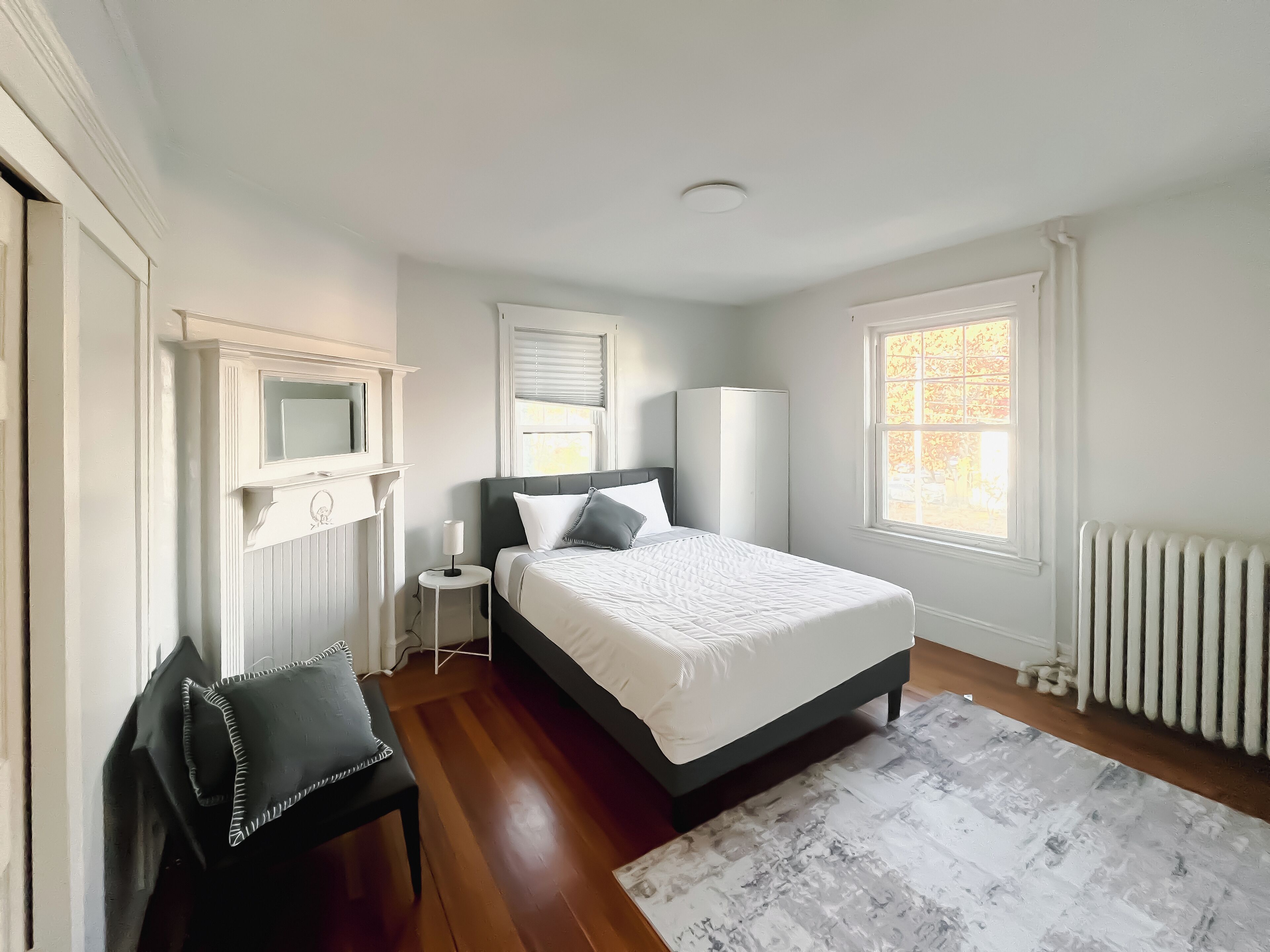 A bedroom with a bed, a chair, a nightstand, a radiator, and a window.