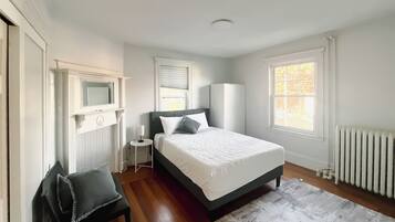 A bedroom with a bed, a chair, a nightstand, a radiator, and a window.