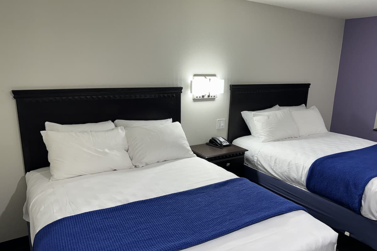 Two double beds with white linens and blue bedspreads, a nightstand with a phone, and a wall-mounted light fixture.