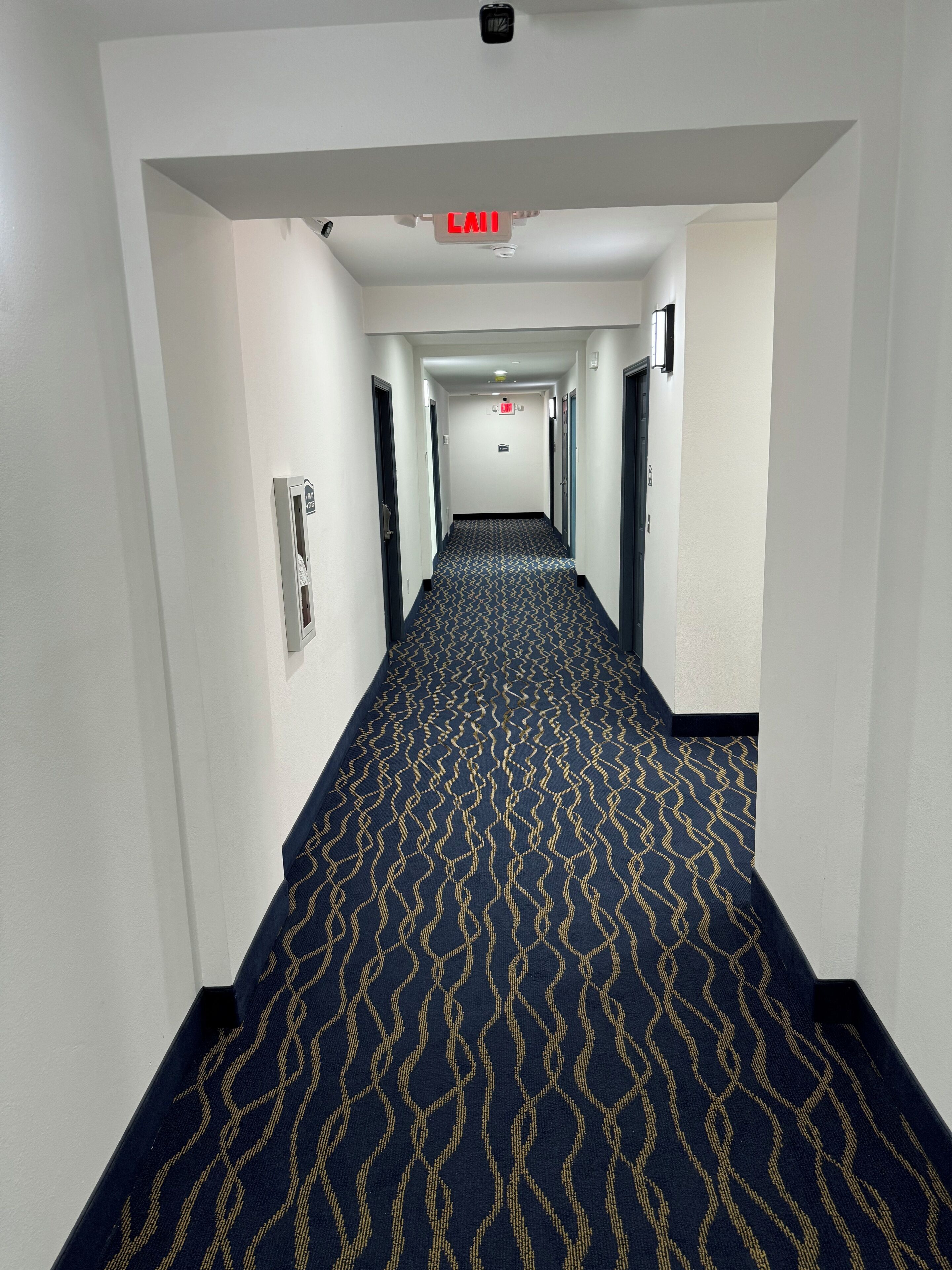 Un couloir d’hôtel avec un tapis à motifs, des portes des deux côtés et un panneau de signalisation indiquant la sortie au plafond.