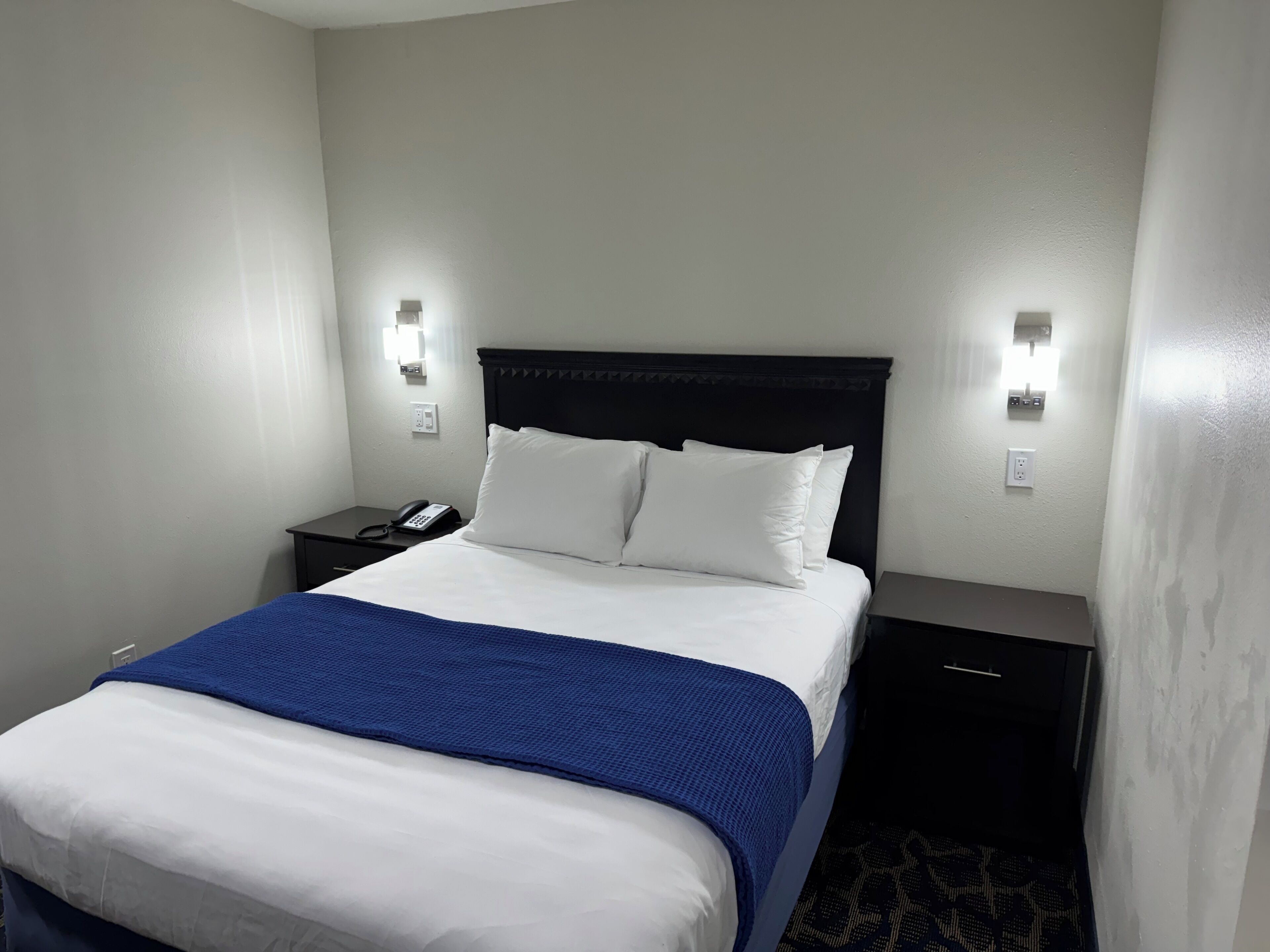 Habitación de hotel con cama, mesita de noche con teléfono y luces fijadas a la pared.