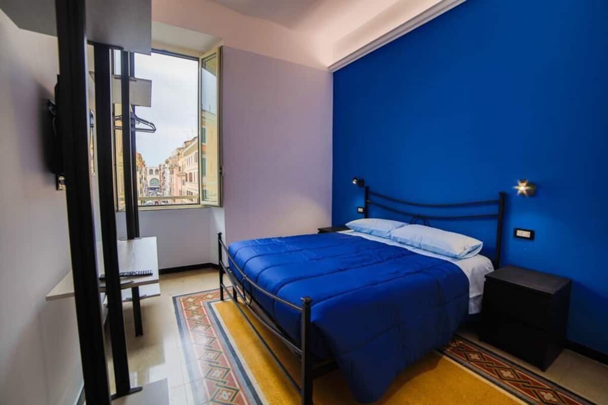 A bedroom with a bed, a nightstand, a desk, and a window with a view of buildings.