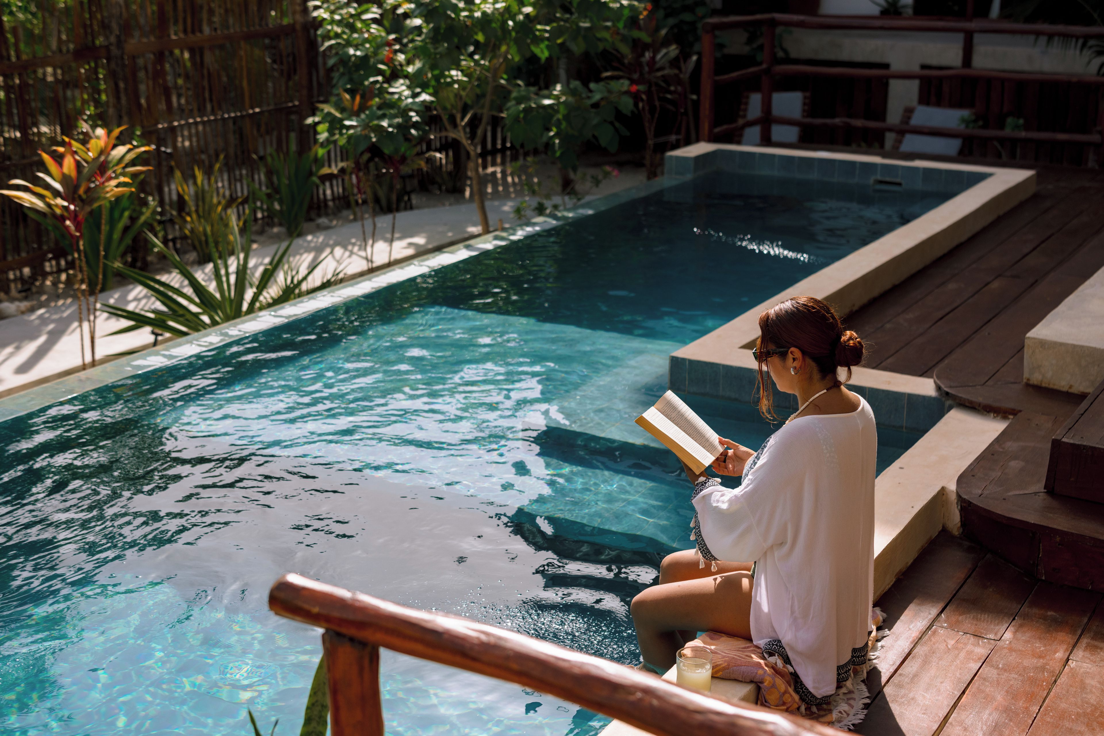 A person reading by a pool with a wooden deck and a bamboo fence in the background.
