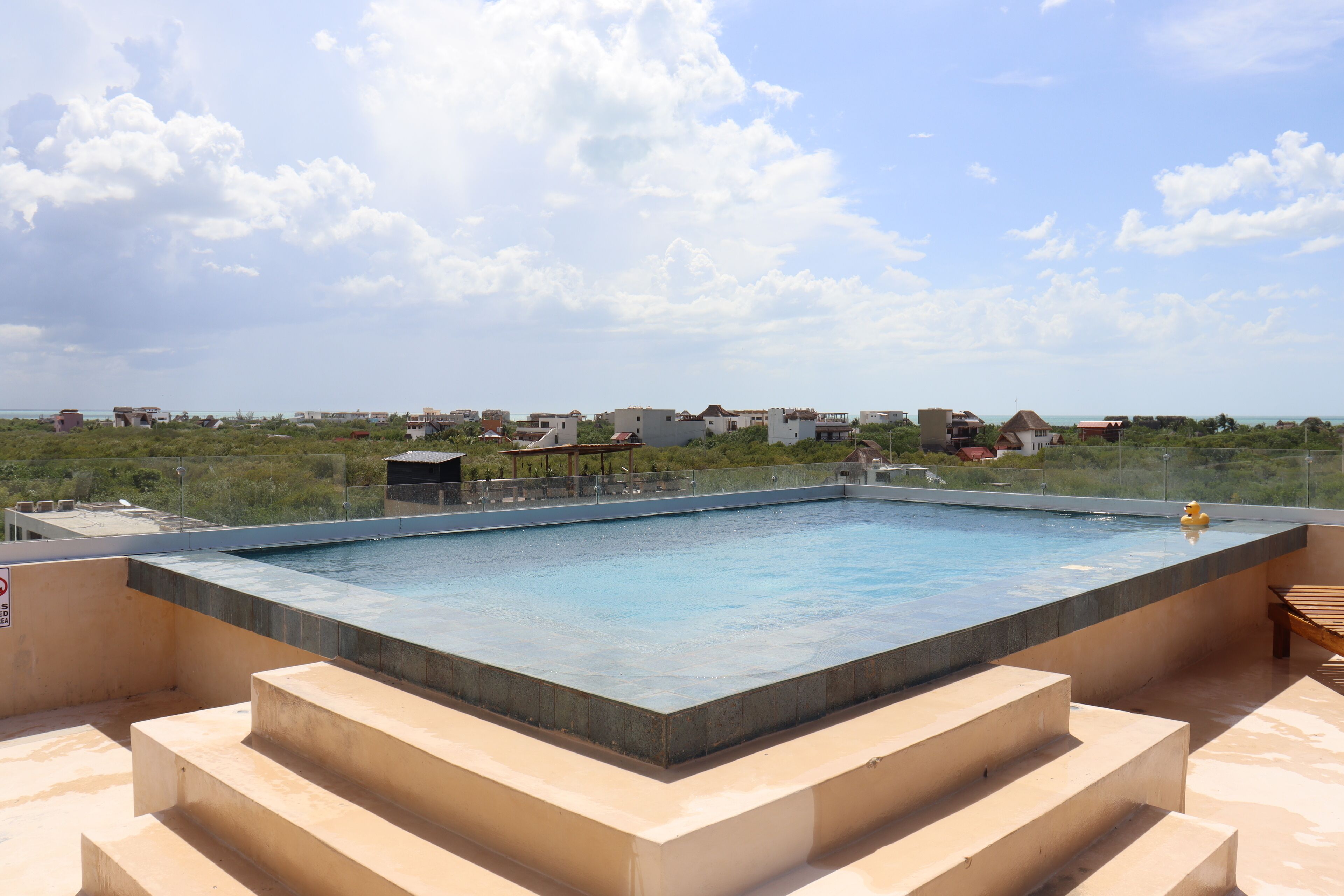 A rectangular swimming pool with a wooden deck and steps leading up to it, surrounded by a clear sky and distant buildings.