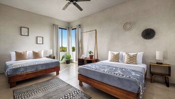A hotel room with two beds, a ceiling fan, a large window with curtains, a potted plant, and a small table with a lamp.