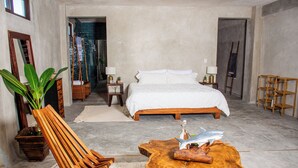 A bedroom with a bed, wooden side tables, a chair, a coffee table, and a large plant. - LaFamilia Casa and Suites (Isla Holbox)