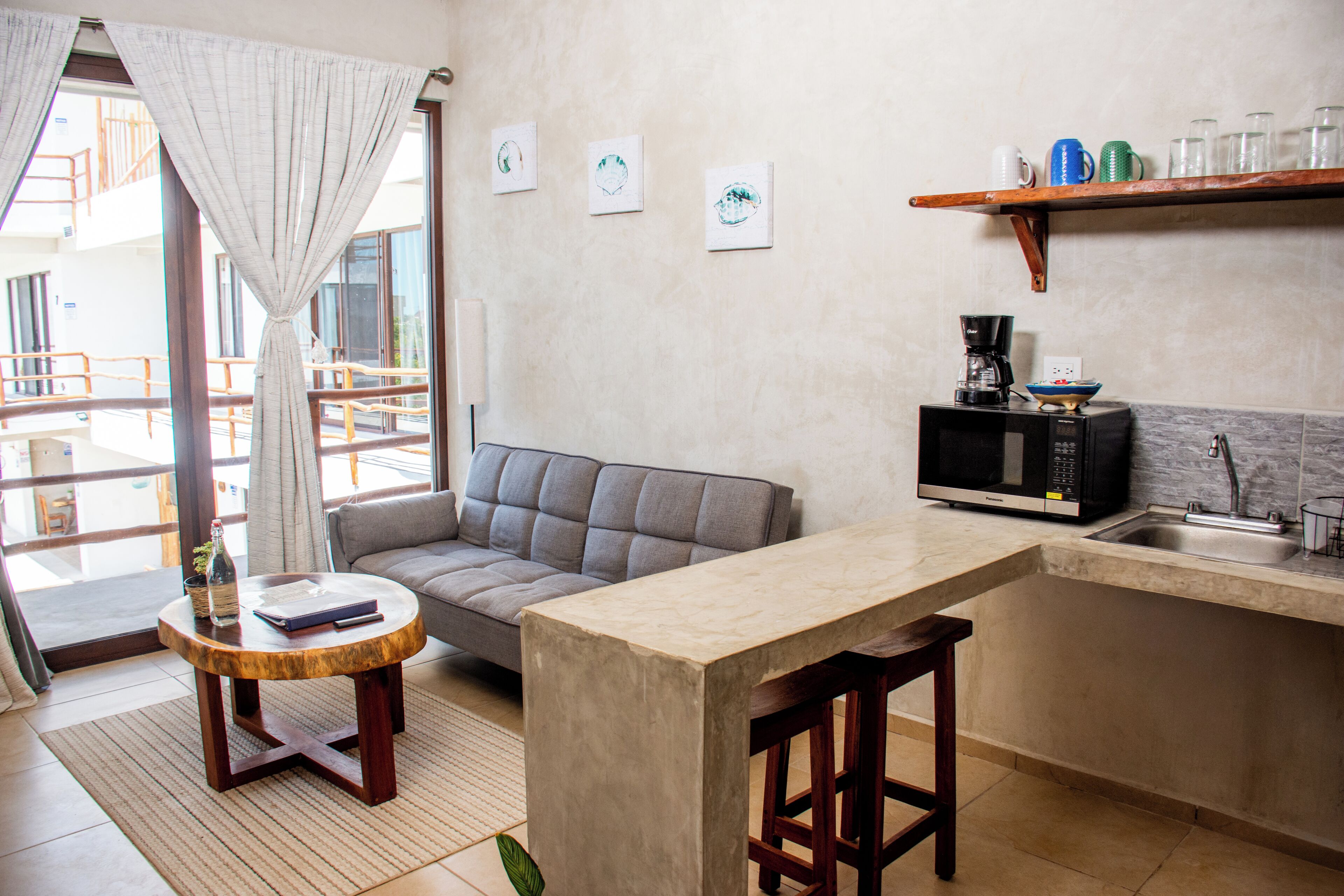 Una cocina moderna con un sofá gris, una mesa redonda de madera y un taburete pequeño de bar. Hay tres cuadros en la pared y una cafetera sobre la encimera.
