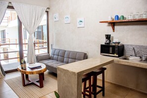 A modern kitchen with a grey sofa, a round wooden table, and a small bar stool. There are three framed artworks on the wall and a coffee machine on the counter. - LaFamilia Casa and Suites (Isla Holbox)