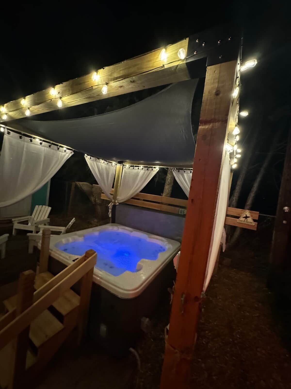 A hot tub under a canopy with string lights, surrounded by wooden chairs and a wooden structure.