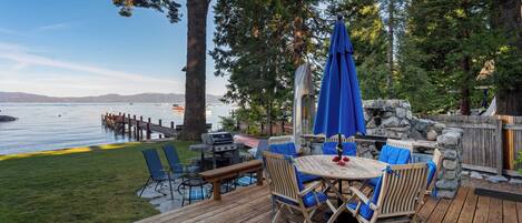 A wooden deck with a table and chairs, overlooking a lake with boats and a dock.