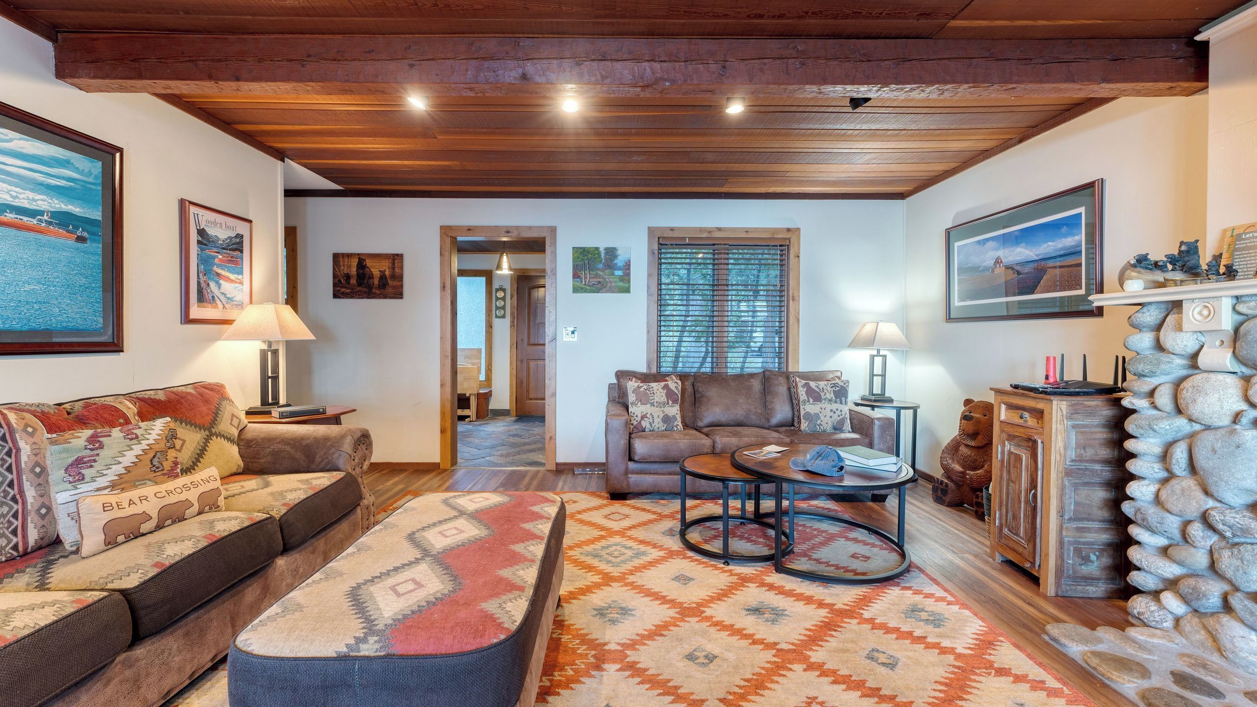 A cozy living room with a wooden ceiling, a stone fireplace, and a view of the dining area.