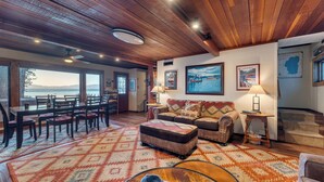 A cozy living room with a wooden ceiling, a large window overlooking a body of water, a dining area with a table and chairs, and a sofa with a patterned throw. - Lakefront Haven by Avantstay Minutes to Ski Resorts Private Dock (Homewood)