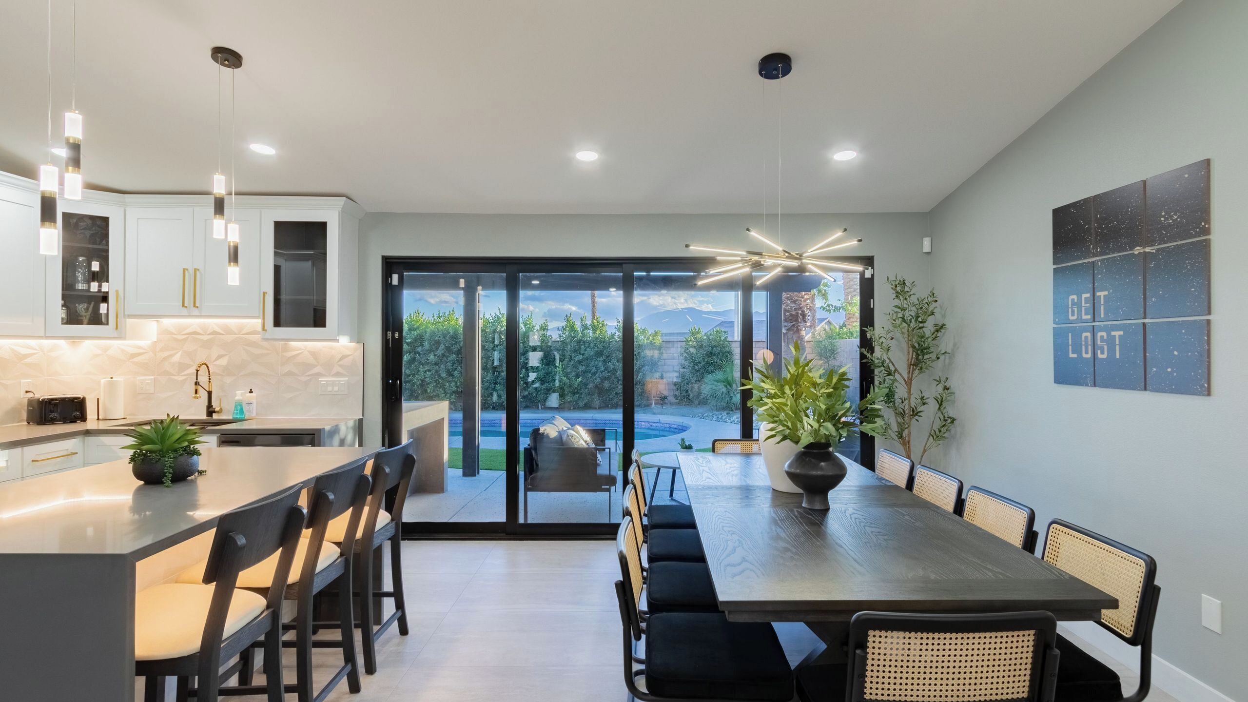 Une cuisine moderne dotée d’un îlot central, d’un coin repas avec des chaises, et offrant une vue sur une piscine et un jardin grâce à des portes vitrées.