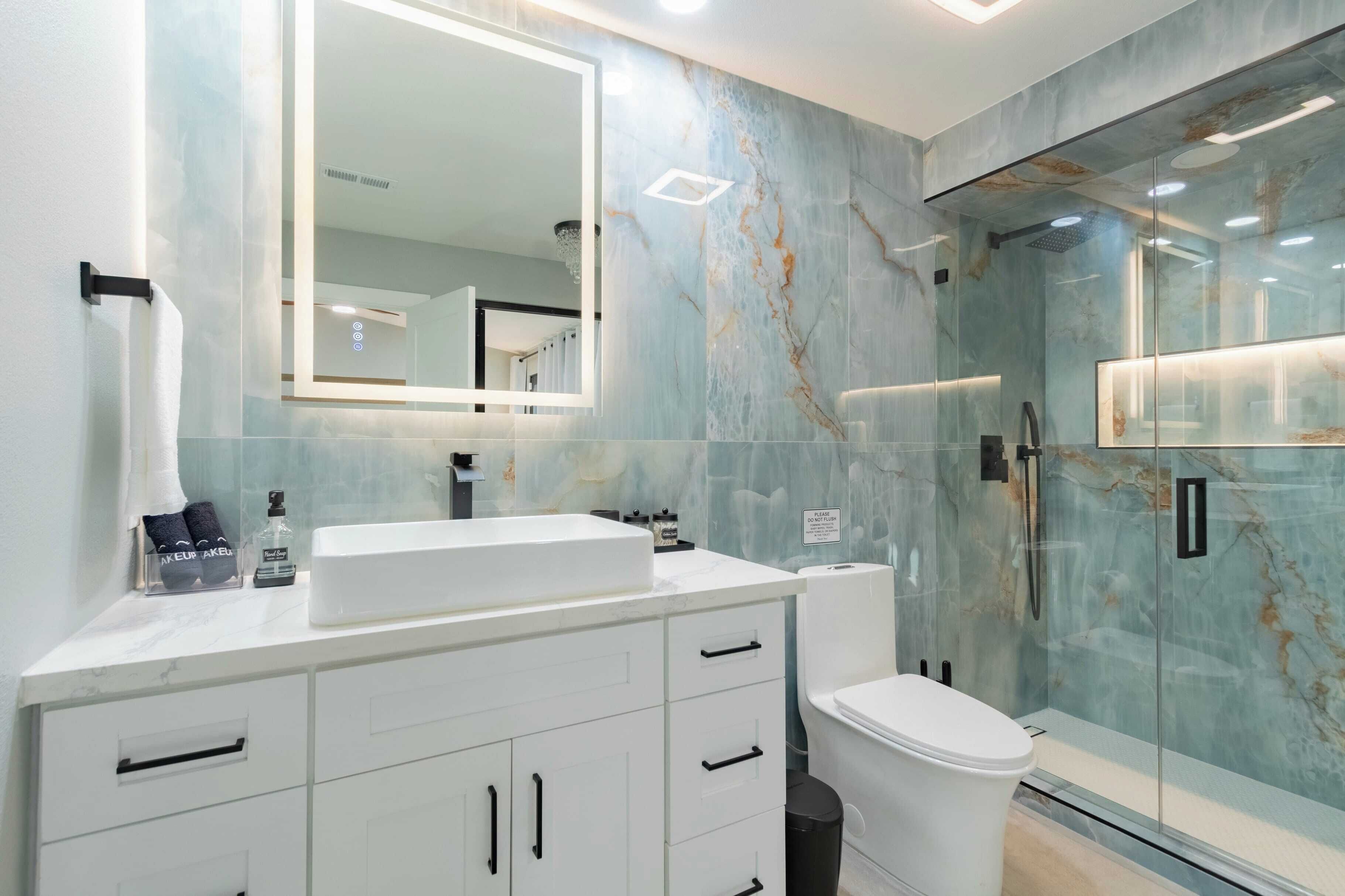 A modern bathroom with a marble vanity, a large mirror, a glass shower enclosure, and a toilet.
