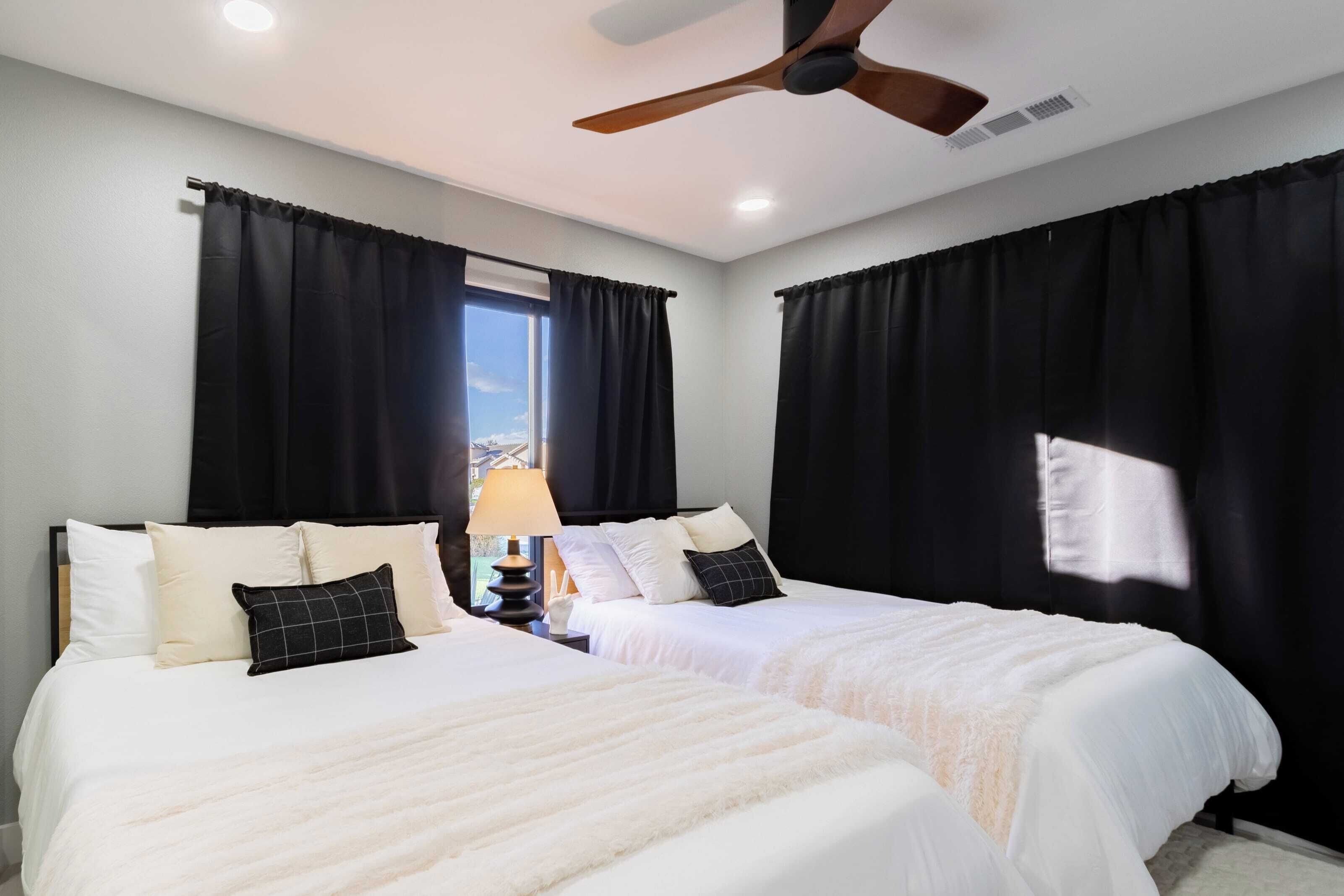 A hotel room with two beds, black curtains, a ceiling fan, and a window with natural light.