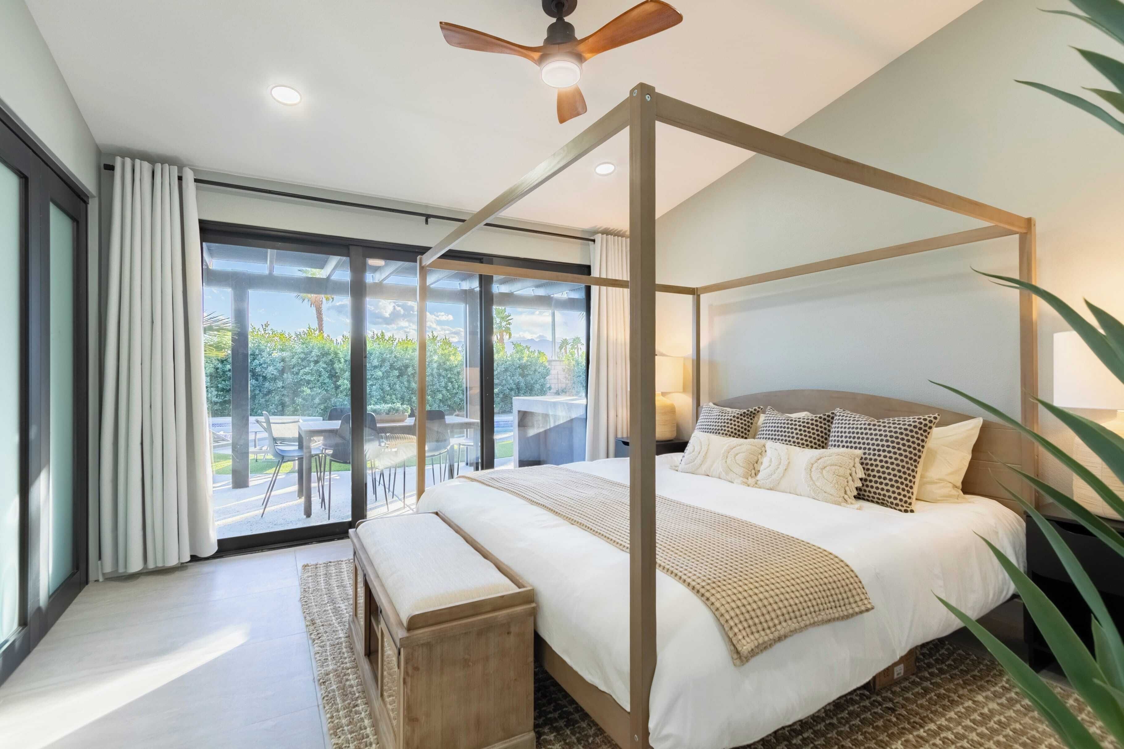 Un dormitorio moderno con cama con dosel, ventilador de techo y vistas al exterior a través de amplios ventanales.