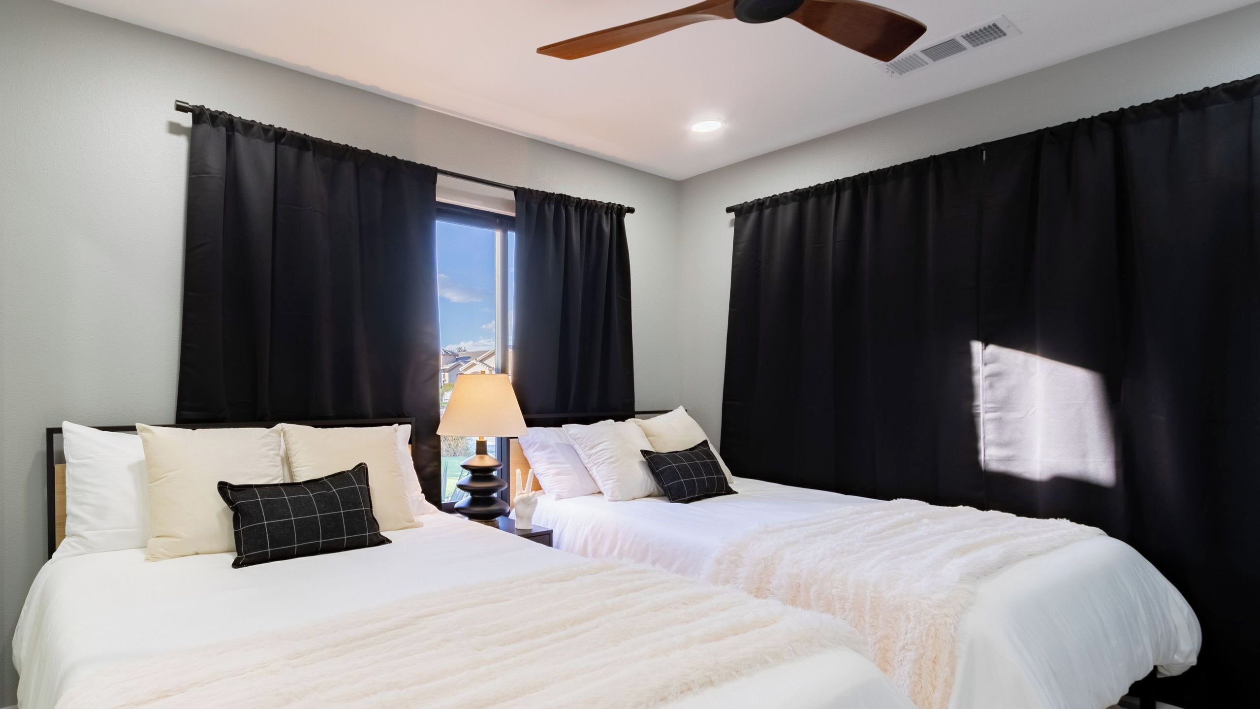A hotel room with two beds, a ceiling fan, a window with curtains, and a nightstand with a lamp.